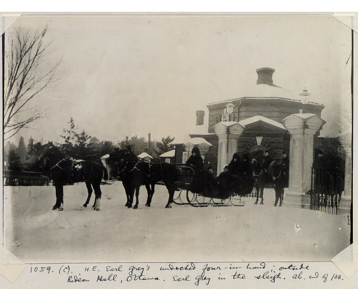 A winter scene near a gate there is a four-horse drawn sleigh and other men on horses behind it A winter scene near a gate there is a four-horse drawn sleigh and other men on horses behind it
