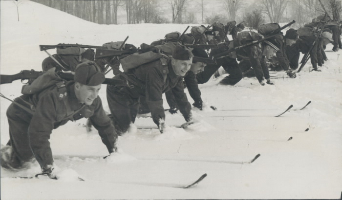 A large number of men on skis kneeling on the snow smiling in uniforms