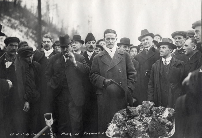 Men in formal attirecircled around a men with wheelbarrow full of dirt