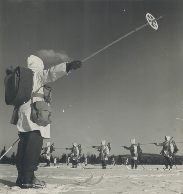 Men in white uniforms on skis holding up ski poles