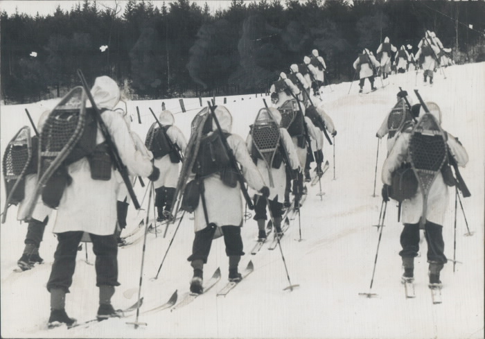 A troop  all in white coats  on skis with snow shoes and rifles