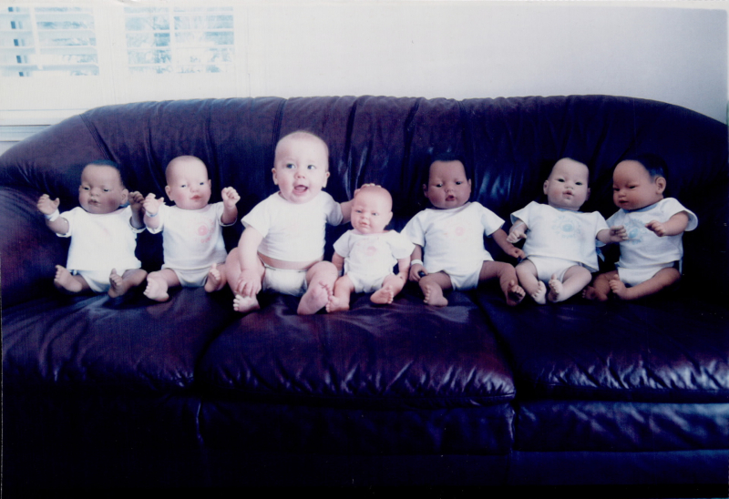 Baby dolls and a baby sitting on a couch.
