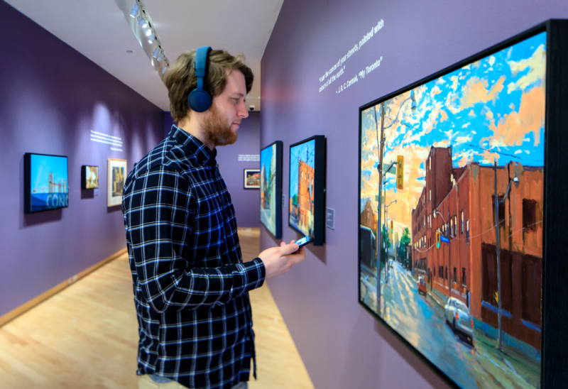 Man wearing headphones and holding phone looking at painting in the TD Gallery Man wearing headphones and holding phone looking at painting in the TD Gallery