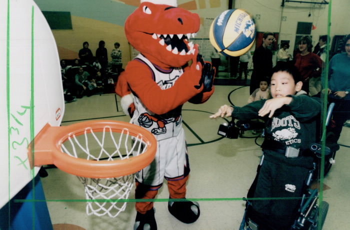 Boy in wheelchair shots a basketball as the raptors mascot claps