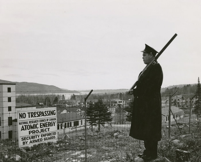 A guard stands watch at the perimeter of the science base. He holds a musket ceremoniously and looks towards the buildings.