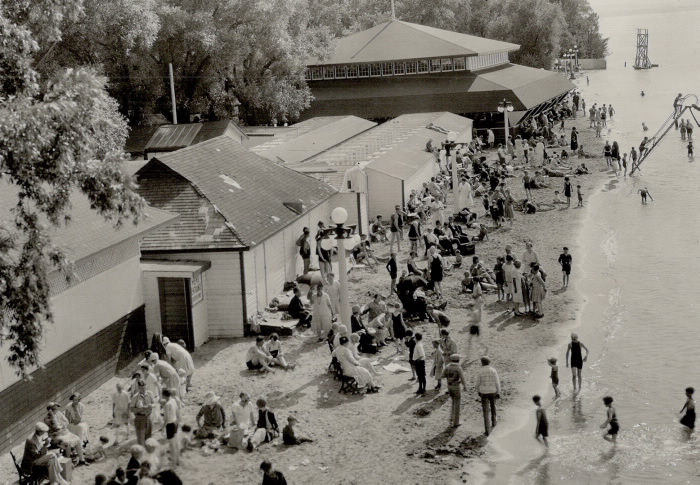 Beach with children in water and adults on beach by building in vintage clothes