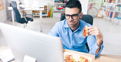 A man eating pizza in front of a computer station.