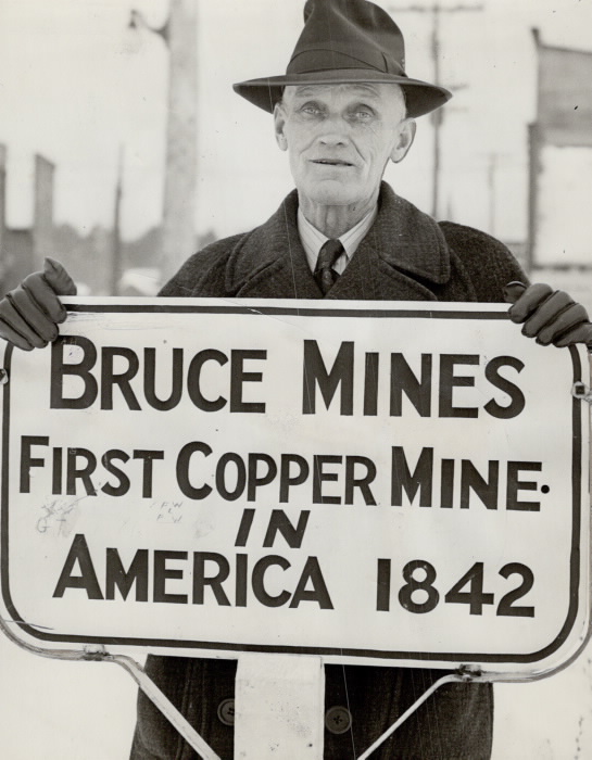 Photo of Albert Grigg, mayor of Bruce Mines in 1943, holding sign that reads "Bruce Mines: First Copper Mine in America 1842".