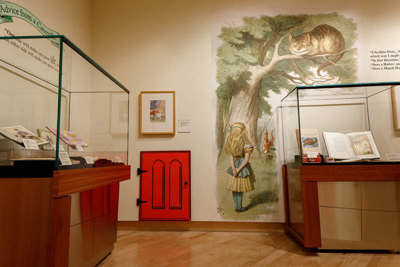 Gallery interior with cases with books and on the wall is an Alice in Wonderland mural as well as a swinging red door