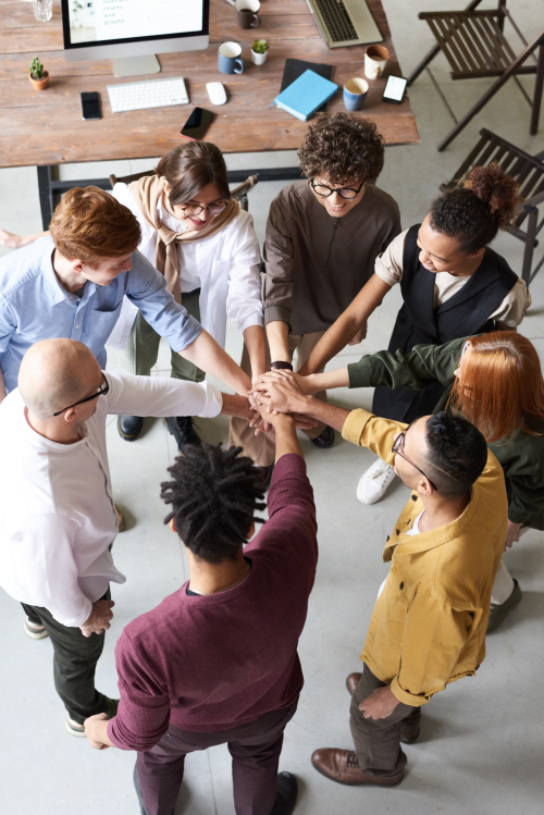 A group of diverse people in an office standing in a circle putting their hands together in the middle. 