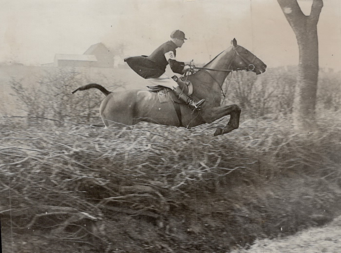 Photo of man riding hours jumping over brush with farm in distant background