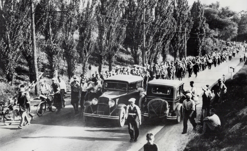 Large group of men walking along street with two cars