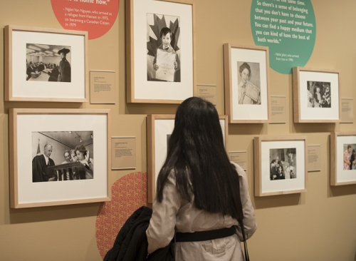 A woman looks at a wall of framed black and white photographs of people of all ages attending citizenship ceremonies. 
