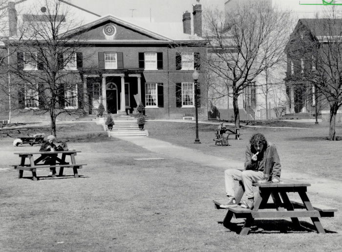 Photo of picnic table with people eating in a park with large brick building in background