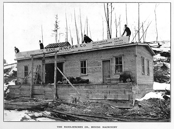 Small shop with banner lifted on roof by men which reads rock drill air compressors pimps pip valves fittings