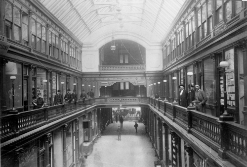 A black and white photograph of the Toronto Arcade. A boy and several men wearing hats stand around the railing on the second floor and look at the camera.