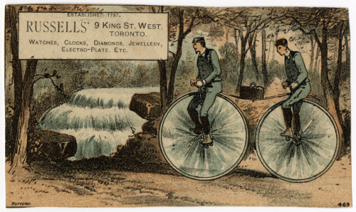 Full colour trade card with two men riding a bicycle with two large wheels and a lunch box sitting between them