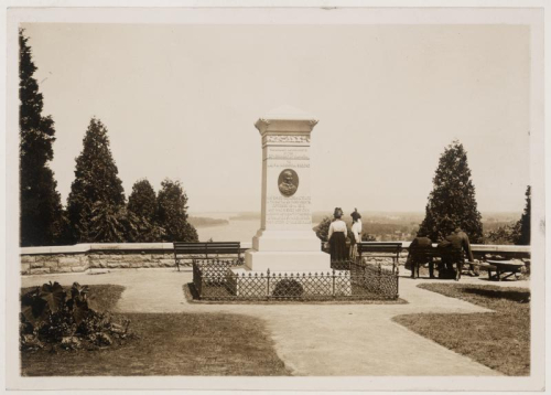 Vintage photo of monument with plaque on it and two bystanders nearby in surrounding garden