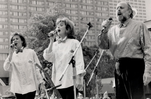 Sharon, Lois, and Bram singing with microphones at Toronto's Nathan Phillips Square