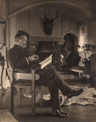 Arthur Conan Doyle is pictured reading and sitting in a chair. A fireplace is in the background.