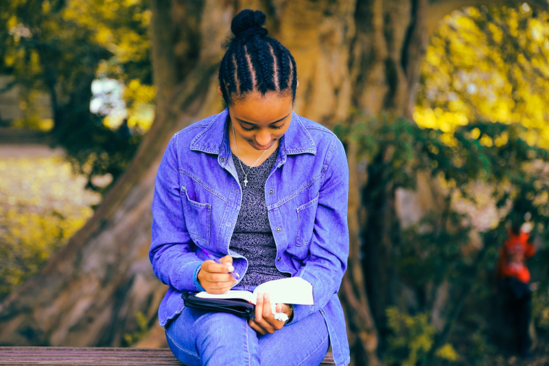 Young woman wearing a denim jacket reading on a bench surrounded by trees