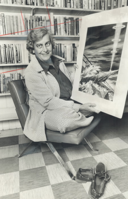 Judith Merril in black and white photo siting cross-legged on chair holding up science fiction illustration