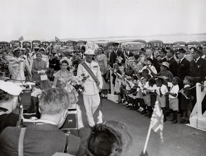The Queen carrying flowers alonside man in uniform surrounded by onlooers waving British flags