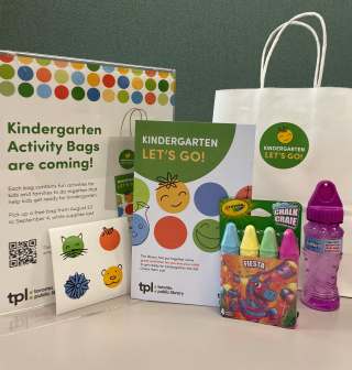 A Kindergarten Let's Go activity booklet, displayed with stickers, a pack of 4 sidewalk chalk, a bottle of bubble solution. A white paper bag with a round green sticker. A sign with text: Kindergarten Activity Bags are coming! A Kindergarten Let's Go activity booklet, displayed with stickers, a pack of 4 sidewalk chalk, a bottle of bubble solution. A white paper bag with a round green sticker. A sign with text: Kindergarten Activity Bags are coming!