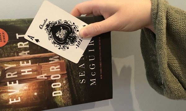 A hand sticking out of a large green sweater sleeve holding a playing card ace of spades and a book