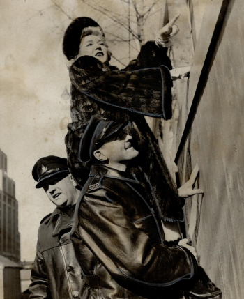 Mary Pickford in fur coat looks over fence supported by two men