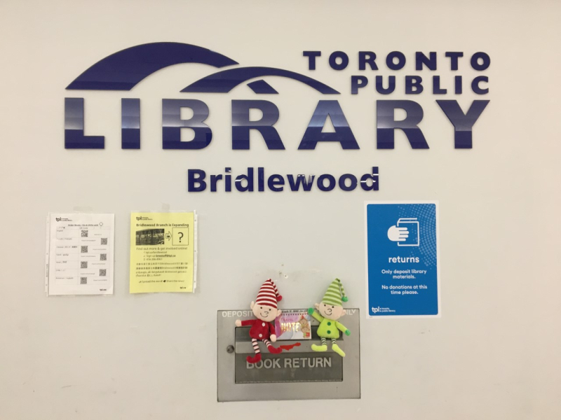 Two stuffed toy elves perched on the edge of a book return slot. The slot is located below a large Toronto Public Library sign and the name of the branch, Bridlewood. There are three posters and flyers on the wall. Two stuffed toy elves perched on the edge of a book return slot. The slot is located below a large Toronto Public Library sign and the name of the branch, Bridlewood. There are three posters and flyers on the wall.