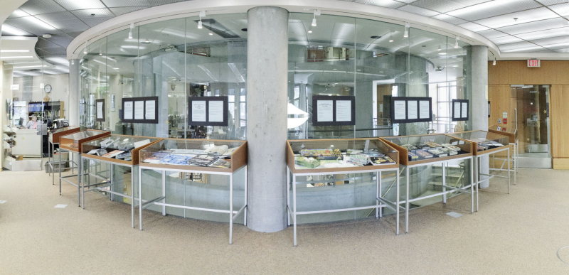 Eight glass display cases are shown in the Merril Reading Room displaying books from the collection. 