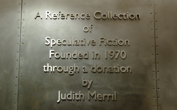 Raised letters appear in a wall of sheet metal from the front of the Merril reading room. Text reads "A Reference Collection of Speculative Fiction Founded in 1970 through a donation by Judith Merril"