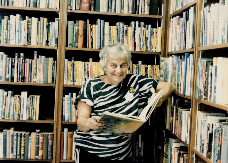 A woman with grey hair and a stripped shirt poses with an open book in front of bookshelves