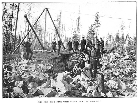 Apparatus on rocks with 10 men standing by looking at camera and subtitle The Red Rock Mine with Steam Drill in Operation
