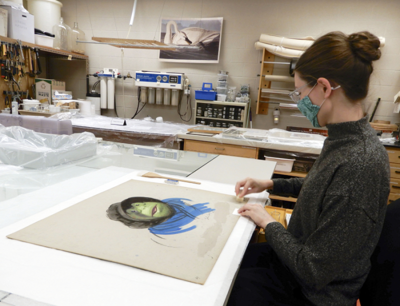A masked conservator is removing dirt from a drawing of Toad with a small piece of chemical sponge.