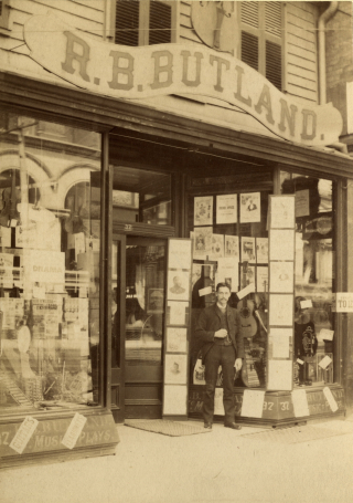 A man with a moustach stands outside a storefront. The sign for the store appears beneath a large violin. Instruments and sheet music can be seen in the window.
