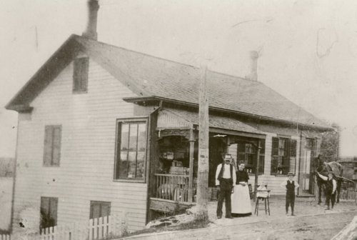 A black and white photograph of a couple a baby and two young children standing in front of a modest house and general store