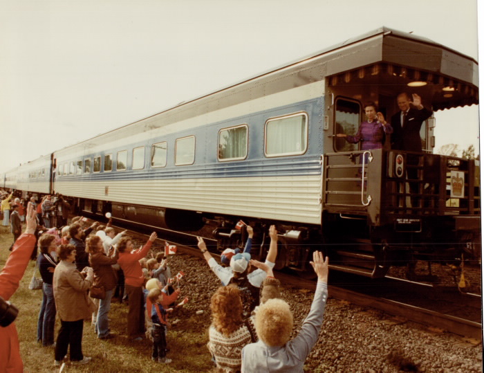 Small crowd with Canadian flag waves on Prince Philip and Queen who are waving from back of passing train Small crowd with Canadian flag waves on Prince Philip and Queen who are waving from back of passing train