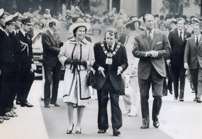 Queen and her husband walk with Mayor David Crombie in street lined with onlookers