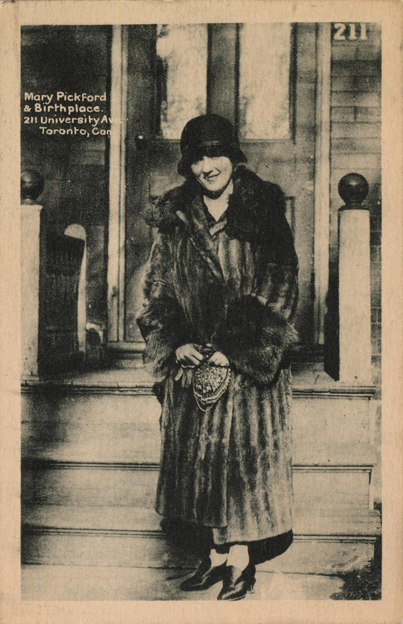 Mary Pickford in fur coat and hat standing in front of the house where she was born.