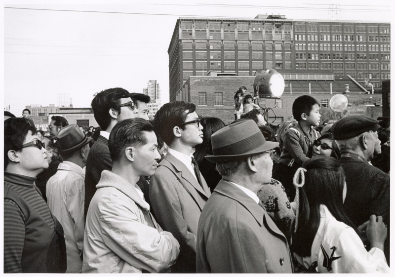 Spectators on street look at event off frame alongside large lights Spectators on street look at event off frame alongside large lights
