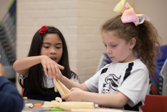 Two children are seated at a table working on a paper craft. One girl is guiding the hand of the other while folding the paper.