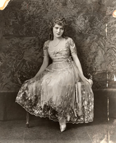 Mary Pickford in elegant drss posing on bench in front of ornate wall