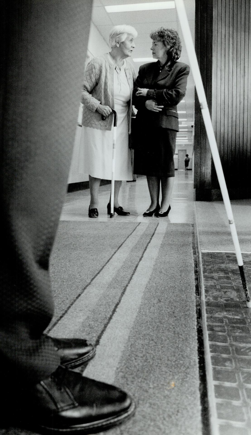 A black and white photo of two women speaking. The younger woman on the right is holding the arm of the older woman on the left. the older woman is holding a cane. In the foreground there is another person's feet and white cane.
