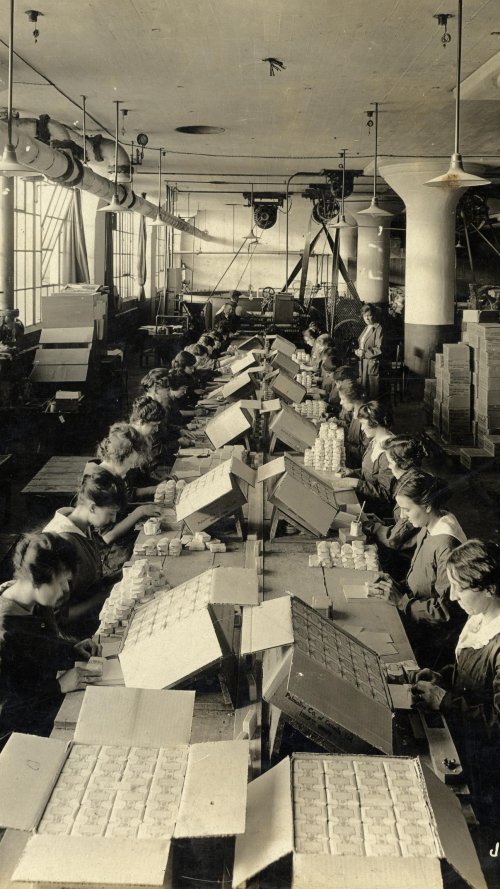 Long table packed with women wrapping bars of soap and big boxes packed with those bars of soap Long table packed with women wrapping bars of soap and big boxes packed with those bars of soap