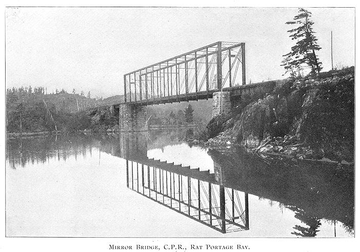 Black and white framed photo of bridge crossing small waterway in an undeveloped wooded area