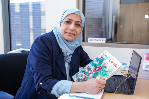 A woman sitting at a desk in front of a laptop and holding a book titles "Coming to Canada". A woman sitting at a desk in front of a laptop and holding a book titles "Coming to Canada".