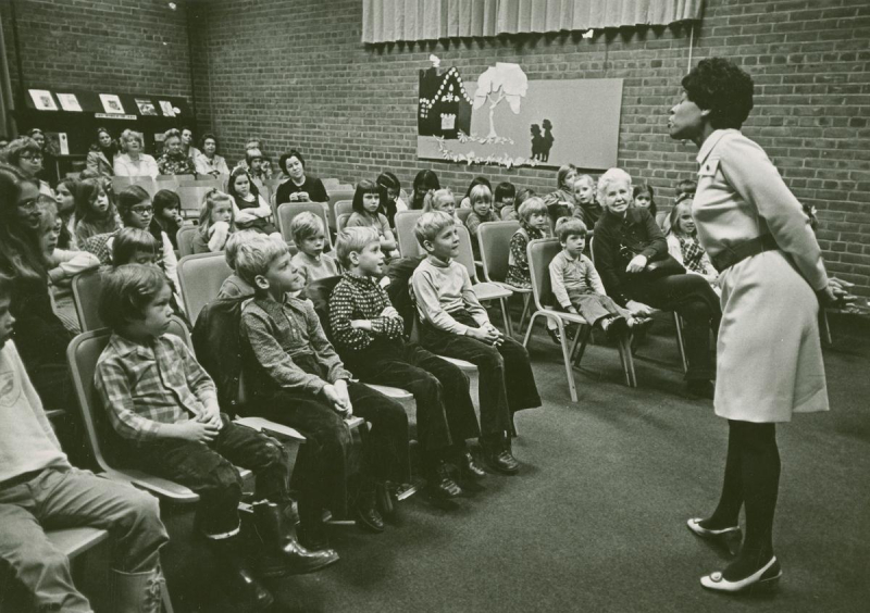 Woman speaking in front of large crowd of children and adults seated theatre style