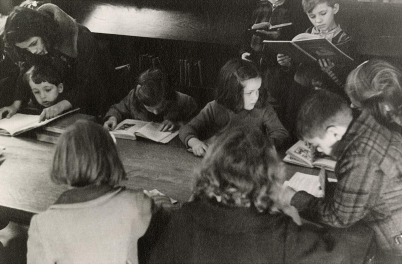 Picture of a group of children seated and standing at a table reading books in a library.